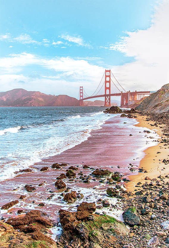 Rocky beach under cloudy blue skies with Golden Gate Bridge in the distance.